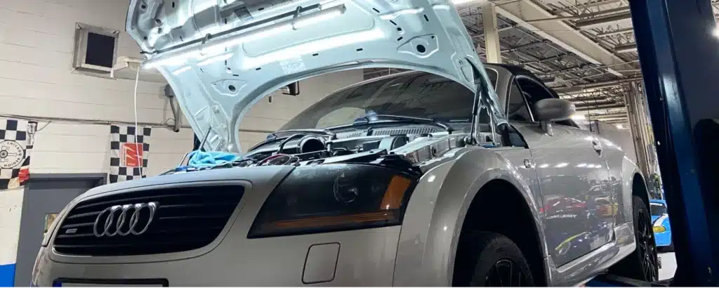 A silver Audi car with its hood open in a garage, being worked on for maintenance or repair.