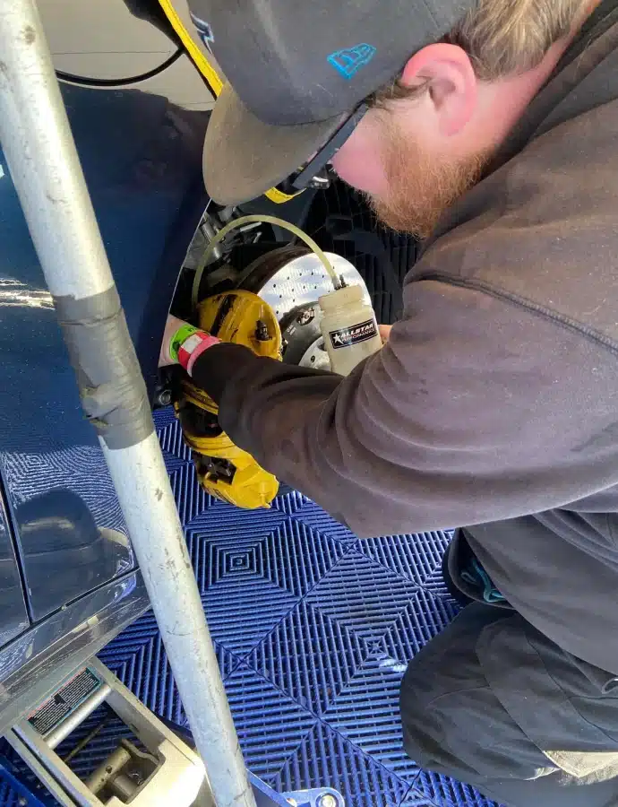 Person working on a car's brake system, wearing a cap and dark clothing, on a blue checkered floor mat.