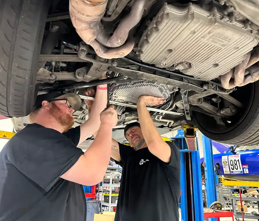 Two mechanics working underneath a lifted vehicle in a garage, focusing on components near the engine area.