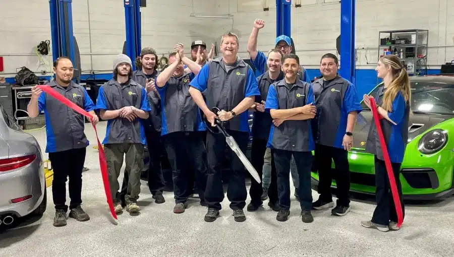 A group of mechanics celebrating a ribbon-cutting ceremony in a garage, with one man holding large scissors in the center.