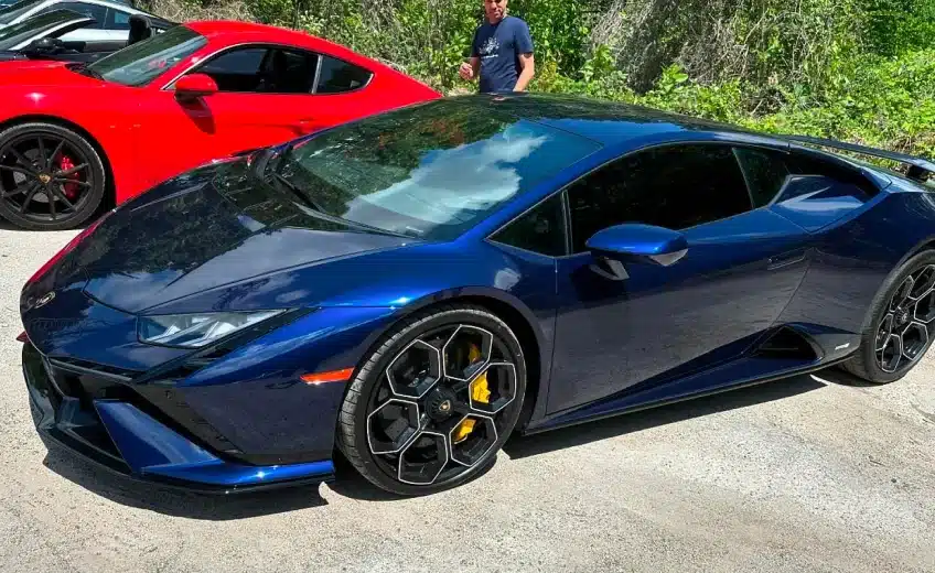 A blue Lamborghini parked on a sunny day with a red sports car and a person in the background.