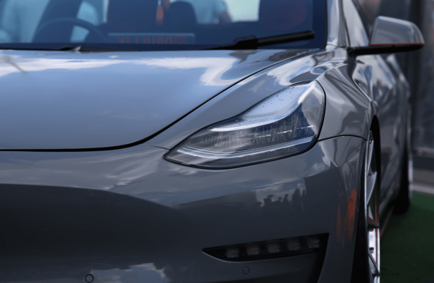 Close-up of a gray Tesla Model 3's front, highlighting its headlight, hood, and side mirror with sky reflections. For Tesla error codes and expert Tesla repair in Gainesville, GA, choose Atlanta Speedwerks.