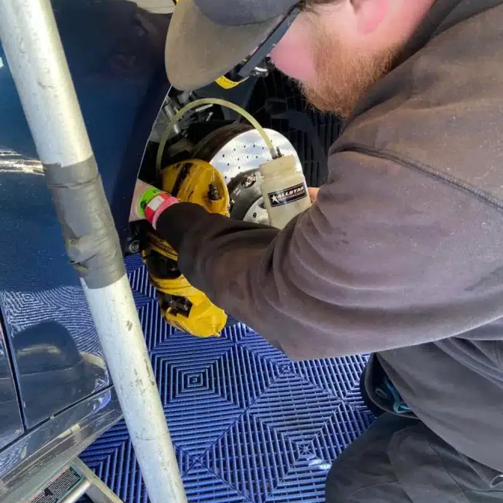 Person working on a car's brake system, wearing a cap and dark clothing, on a blue checkered floor mat.